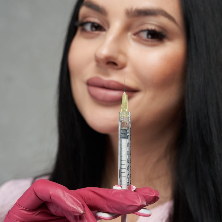 Beautiful female wearing pink gloves showing filled syringe to camera. Close up portrait of attractive brunette woman keeping syringe in front of face and smiling indoors. Concept of beauty care.